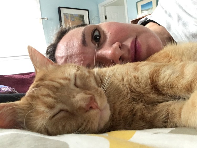 An orange cat is laying on its side with its eyes closed. There is a white woman is laying behind the cat with her eyes open.