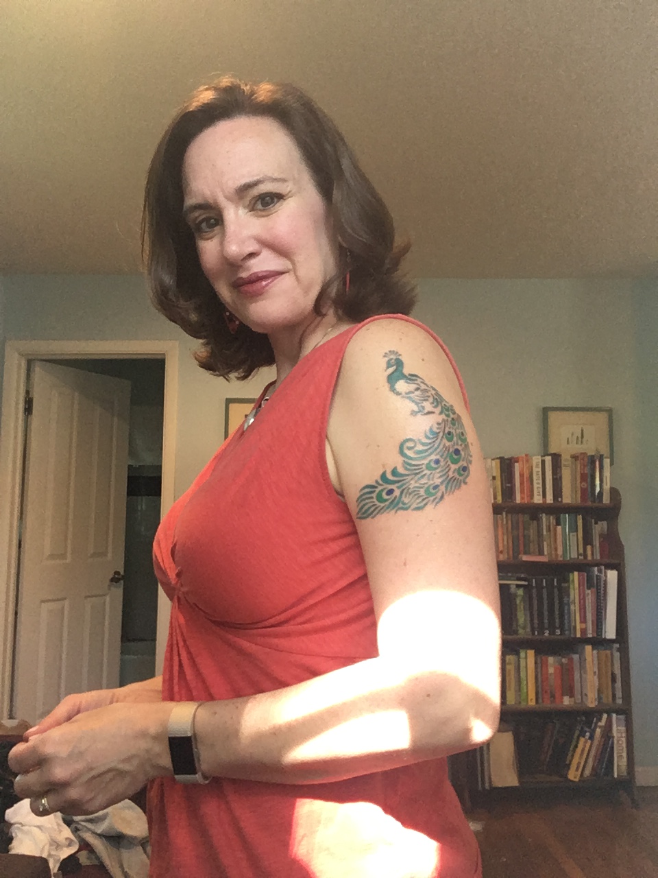 A white woman with brown, shoulder-length hair. She's wearing an orange shirt. Her body faces to the left, she's looking at the camera. On her left shoulder is a colorful peacock tattoo.