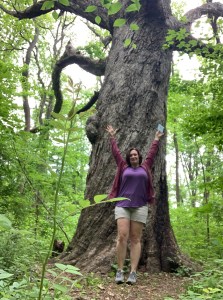 A woman in a purple shirt and shorts, her hands over her head, in front of a very large oak tree. She's in the forest and the leaves are on the trees.