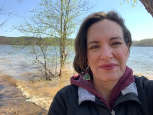 Woman in the foreground with a sweatshirt and coat on. The lake in the background. It's windy with her hair blown and waves on the water. There's a tree with leaves in the water and the other side of the lake is visible.