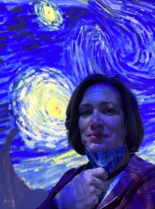 A woman, pulling her mask down, standing in front of a projection on the wall of van Gogh's Starry Night. A bright yellow star is to the left of her face, the background is vibrant blue.