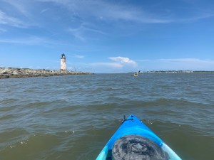 View from the kayak out into the ocean, a small white lighthouse is on the left. It's a bright sunny day, a few whisps of clouds.