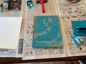 A block with a peacock carving in it, blue ink is spread on the block. There's newspaper on the table underneath, a roller to the right and blank paper to the left.