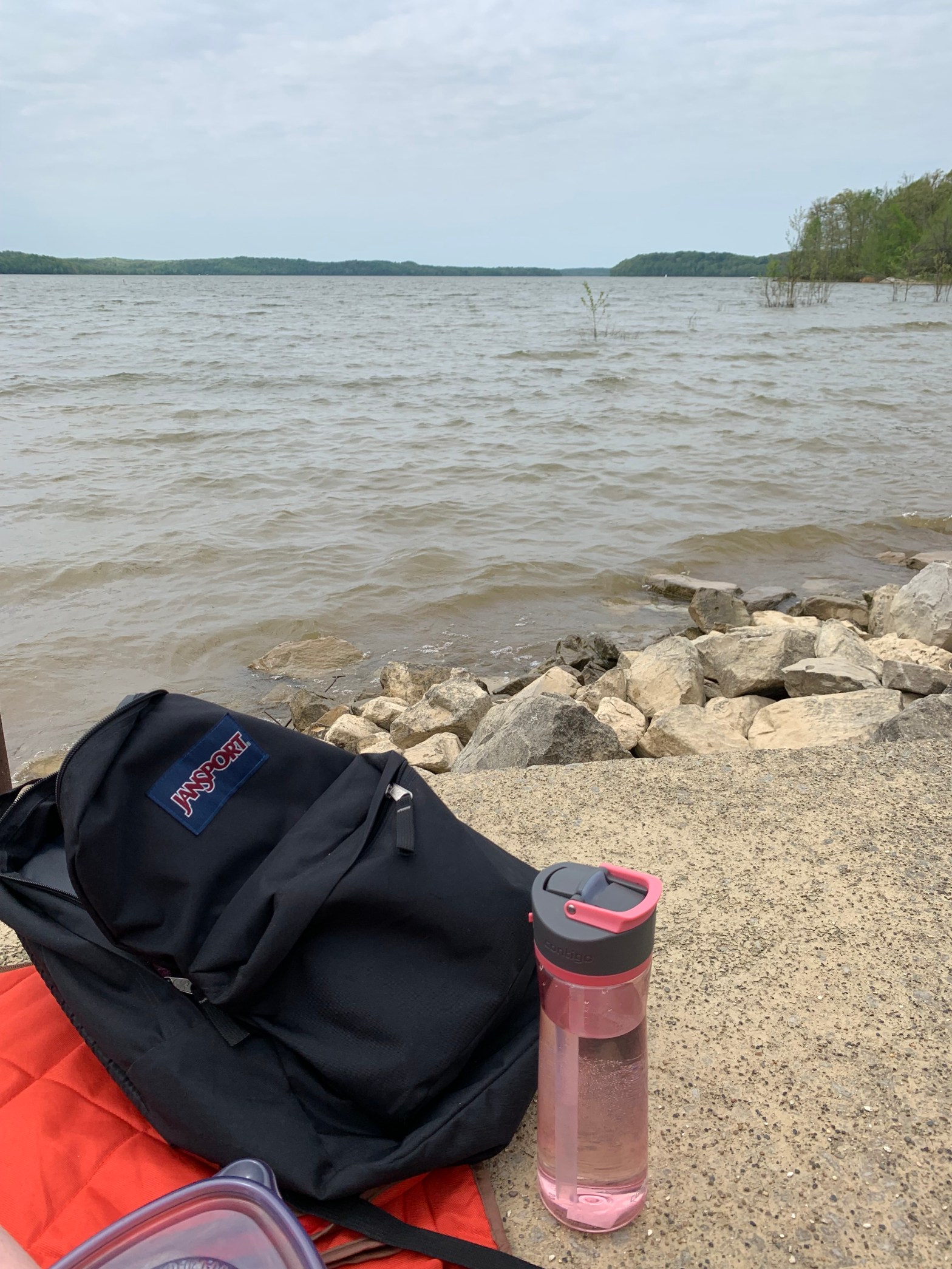 In the foreground, a black backpack, pink water bottle. The lake is in the background. It's a gloomy grey day