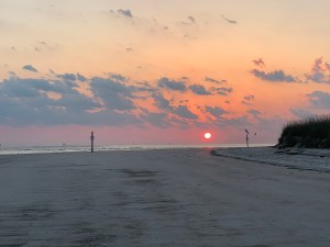beach at the bottom, dune with grass on the right. An orange sun just about the sand. 