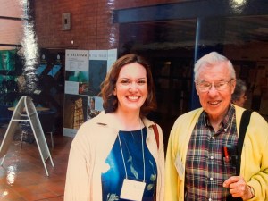 A woman on the left in a blue dress and tan sweater. An older man on the right in a plaid shirt and yellow sweater. They are both smiling. They are both wearing conference name badges. There is a poster board behind them in Swedish that ways, Welcome to Vaxjo University.