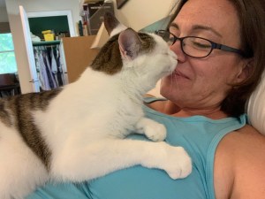 Me, a woman, reclining with a white cat on my chest. We're "booping" noses. I have a blue tank top on and glasses.