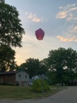 A small pink balloon, lit from underneath, is floating about 12 feet above the ground. There is a neighborhood with two houses in the background. The sky is at sunset, light blue with a few clouds.