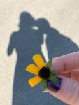 A yellow flower in the foreground held between two fingers. There's dark purple nailpolish on the thumb. In the background are two shadows: on the left a woman with shoulder-length hair and a dress, on the right a slightly shorter shadow
