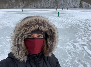 A woman with a red cloth mask on and a hooded coat is in the foreground. A frozen expanse is in the background with some figures way back playing ice hockey. The other side of the lake with bare trees is not far away.