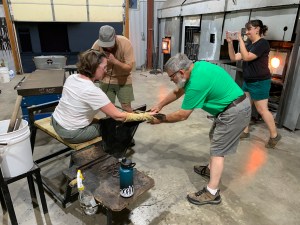 There are four people. A woman is seated, one man is blowing into a glass pipe, another man is holding a paddle to flatten the end of the cup, and a woman is standing near a furnace taking a picture