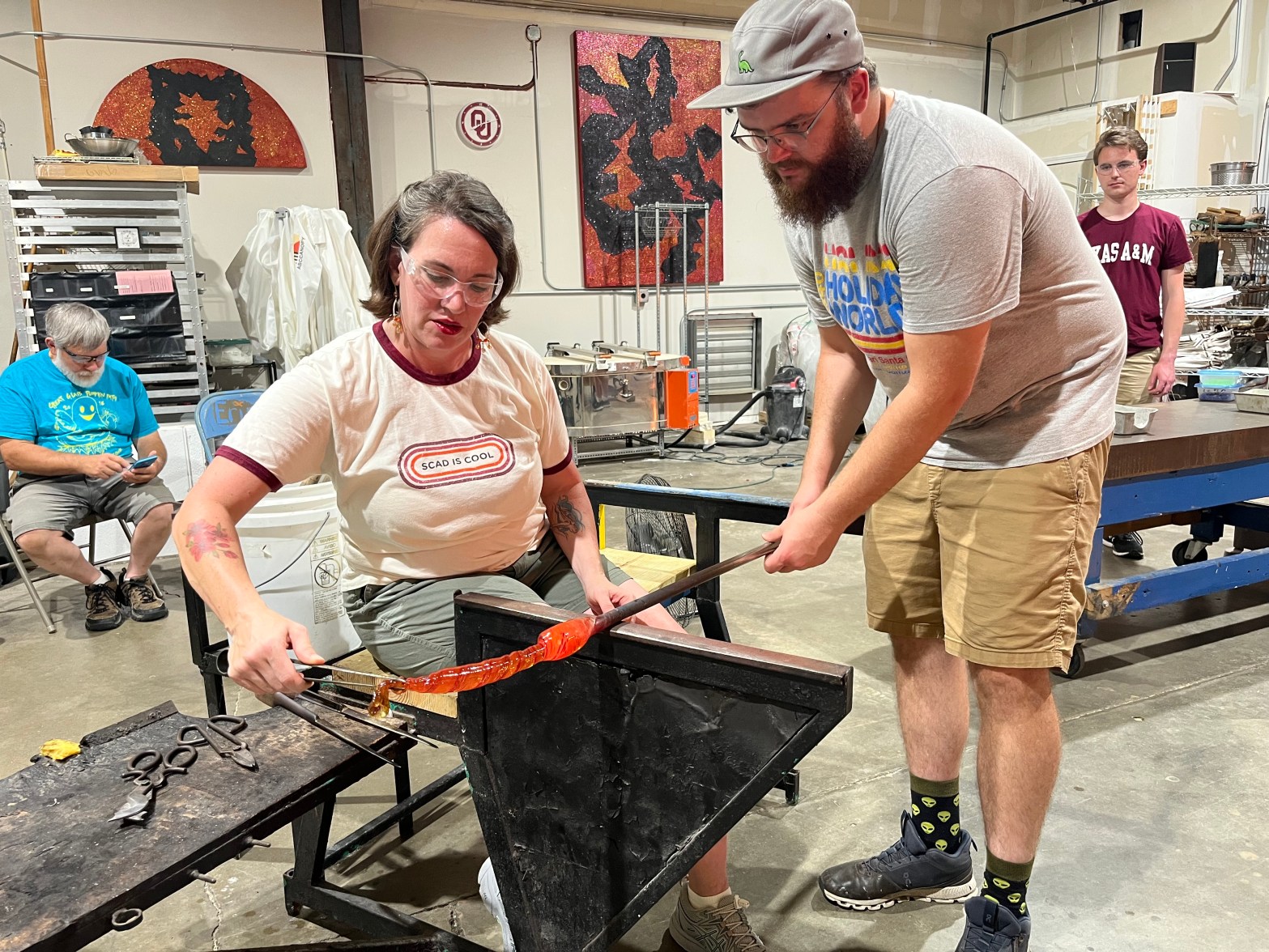 a woman in a t-shirt seated at a bench pulling on a red rod of hot glass. A man in a baseball hat and Holiday World t-shirt is standing next to her. There are tools and benches in the background and two other people behind them.