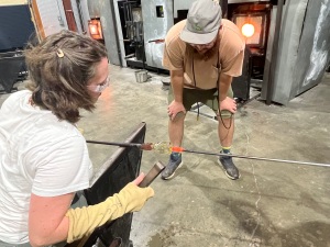 On the left, a woman in a white shirt and brown hair seated. A man is hunched over in the middle of the image. They are both watching two rods with hot glass in between them