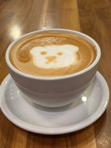 a mug on a saucer filled with caramel colored latte. There's a foam image of a bear on top. The mug sits on a wood table.