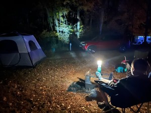 a campsite at night. There's a tent on the left and a red car on the right. The photo is taken from behind someone with a flashlight shining into the woods.