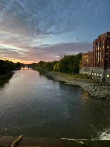 A river with the sunrise in the background. There's a building on the right with lights on and a bridge over the water in the distance.