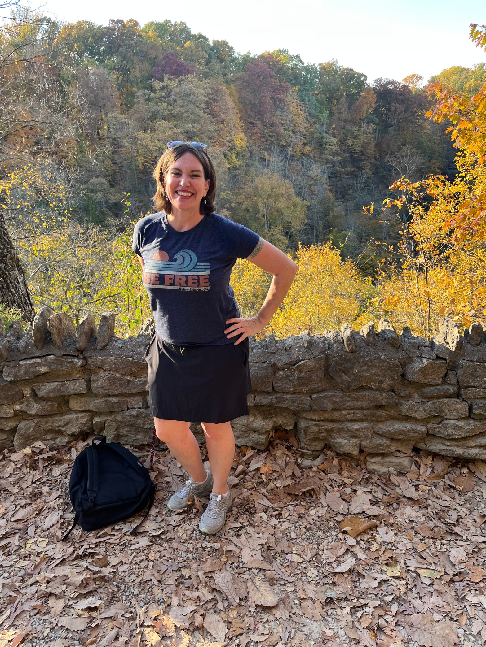 a woman in shorts and t-shirt standing in front of a short stone wall. There's a canyon behind her and colorful trees. There's a book bag at her feet.
