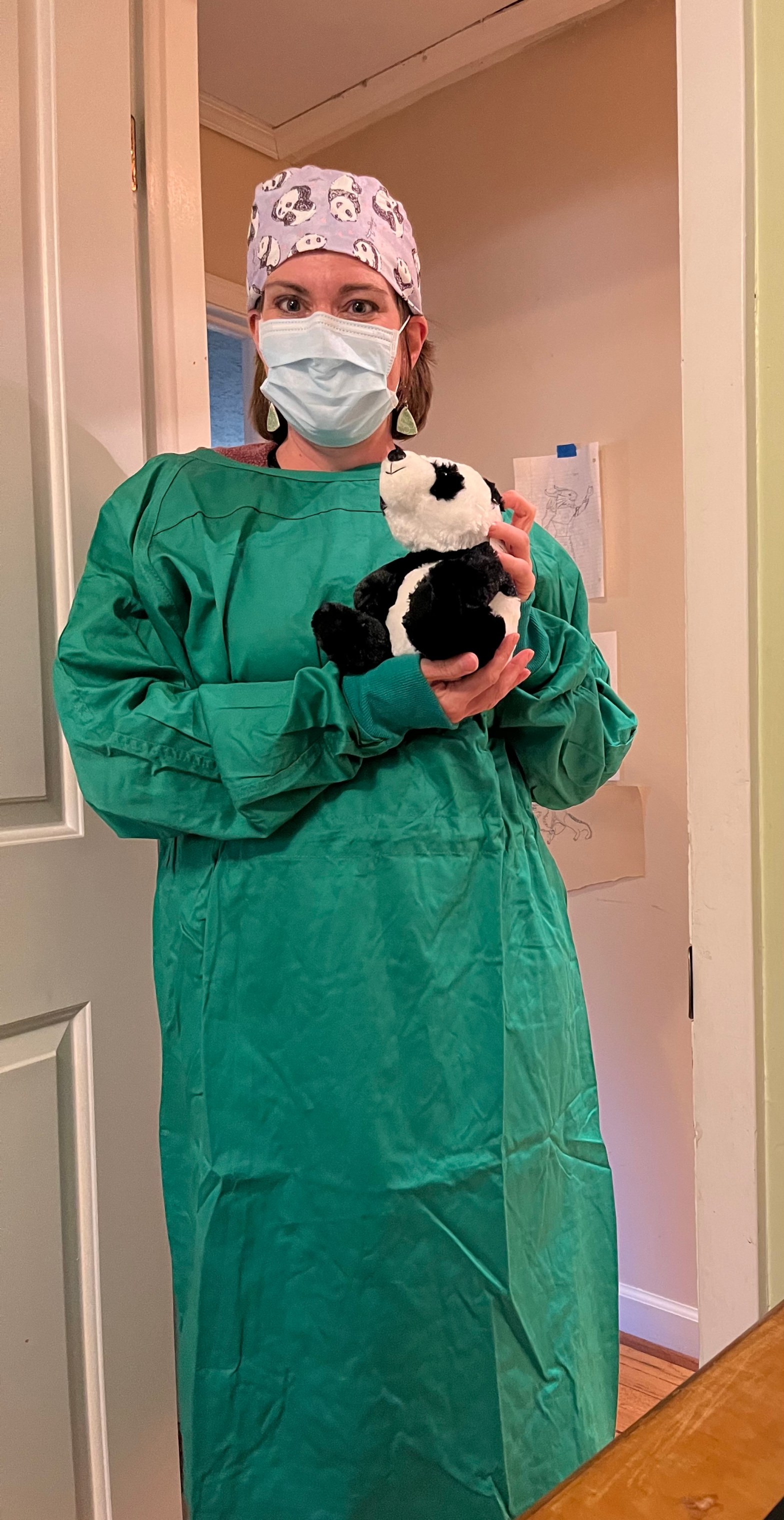 A woman with a green surgical gown on holding a stuffed panda. She has a paper mask and a purple surgeon's hat with pandas on it. She's standing in a doorway.
