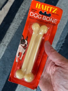 A nylon dog bone still in its wrapper. the cardboard backing is red and says "Hartz" on it. There's a Boston terrier on it. The package is held by a hand on the right and there's a blue carpet in the background.