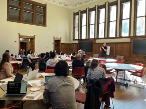 A speaker is at the front of the room, the picture is taken from the back of a conference room. People are seated at round tables. There is a large row of tall windows behind the speaker and wood paneling along the walls