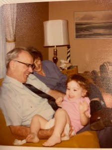 There are three people on a couch. On the left is an older man in a tie and behind him an older woman in a blue dress. They are holding a toddler in a pink dress. They are all smiling and laughing. There's a lamp in the background, a lighthouse table statue, and a painting of ocean waves.