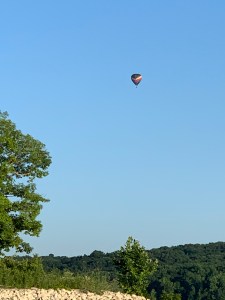 A hot air balloon high over a green hill. There is gravel and a tree in the foreground. The sky is blue and it is daytime. The balloon is black with a diagonal rainbow stripe.