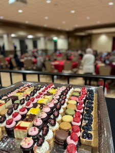 In the foreground, a silver platter of colorful, bitesized pastries. In the blurry background, a conference room of tables in rounds.