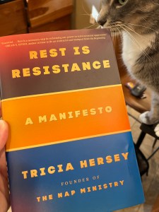 Book cover to Rest is Resistance: A Manifesto, by Tricia Hersey. Each line is in a horizontal color band of red, orange, and blue. A left hand is holding the book. A grey cat is in the top right of the image.