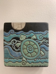 A black tile with a blue and green design of a turtle in the sea. There is a moonlight on the top of the tile.