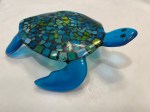 A translucent glass turtle on a white background. The legs elevate the turtle off the surface and the head is raised. The shell of the turtle is mosaic glass - blue, green, yellow tiles.