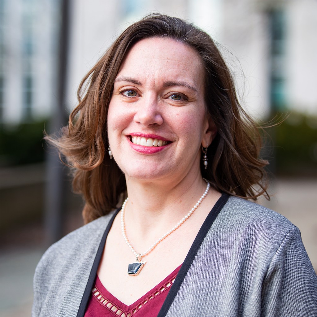 A closeup of a white woman's face. She is smiling gently. She has brown hair that is blowing in the breeze. She is wearing a grey sweater over a red shirt.