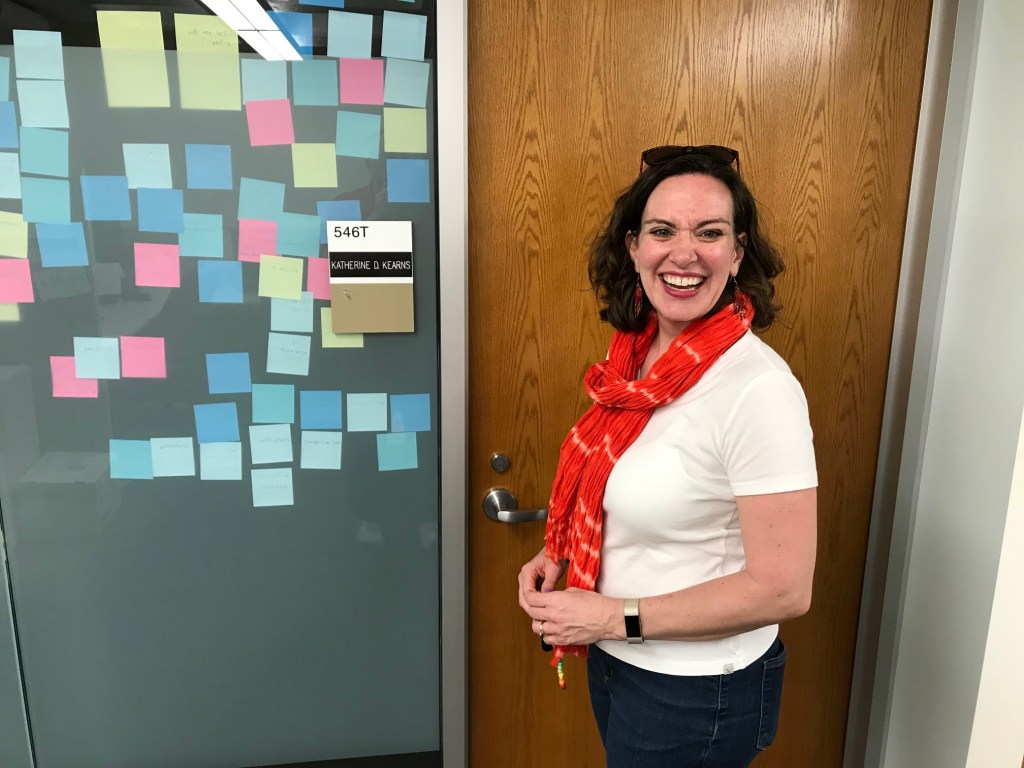 A white woman with shoulder length brown hair and a red scarf is standing in front of a wooden office door. She has a big smile on her face. To the left of the image is a glass wall with colorful notes all over it.