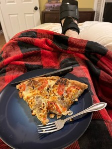 in the foreground, a blue plate with pizza on it. It rests on the lap of someone with a red fleece blanket. Their right foot is extended beyond the blanket; there's a black boot on their foot.