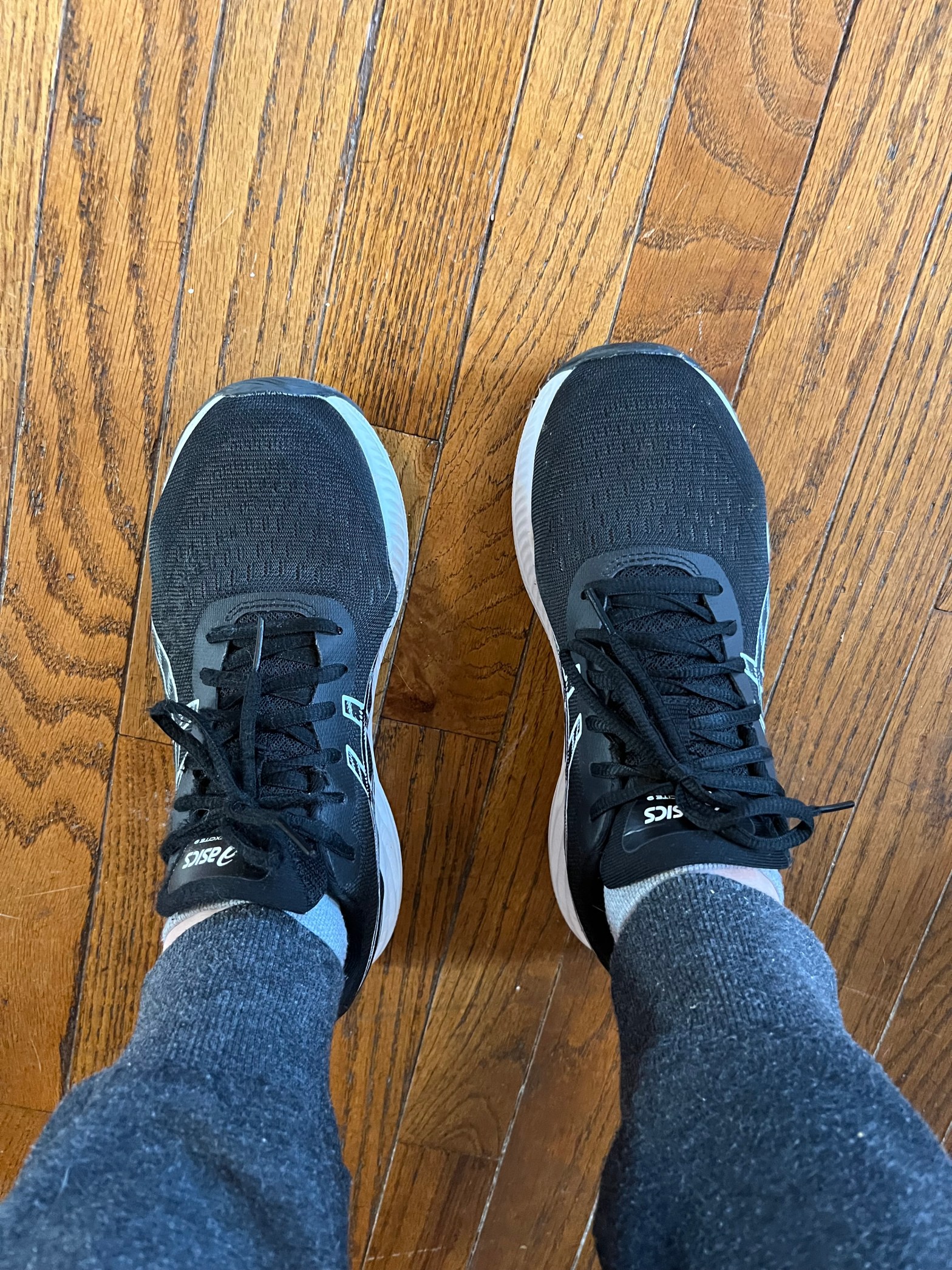 Two feet from mid-calf down. They are wearing black sneakers with white soles. The person is wearing dark grey sweatpants. They are standing on a wood floor.