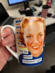 A hand is holding a mug that says "stop me before I volunteer again." There's a woman's face on the mug. The design has 1950s styling.