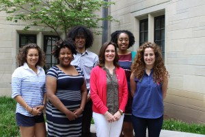 Six people are standing in a courtyard smiling. The trees and plants behind them are green.