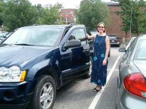 A woman in a long blue dress and heels is standing next to a blue Jeep Wrangler in a campus surface parking lot. The trees are green behind her. She has sunglasses on.