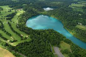An overhead view of Green Lake. It is a comma shape, round with a tail, and is teal in color. It is surrounded by some woods, there is a golf course to the left.