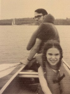 Two people are in a canoe. A girl about 7 is facing the camera, she has brown braided pigtails. A man is facing the other way with his head looking at the camera. There is a sailboat in the background. The picture has a yellow-tinge to it.