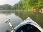 The view from a kayak. The bow and an oar are in the foreground, the lake and the bank with trees is ahead.