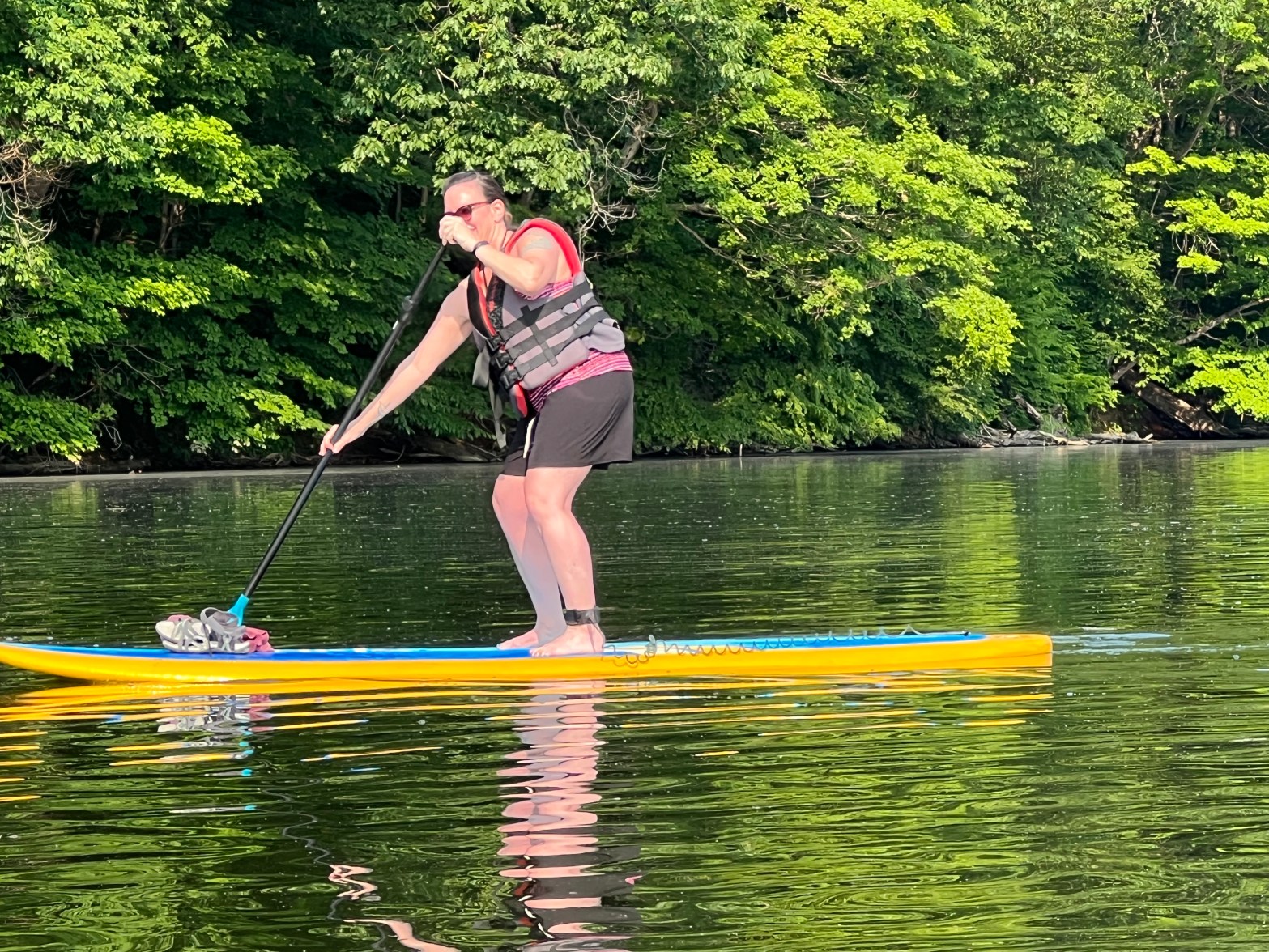 A woman in black board shorts and a life jacket is standing on a paddleboard. She is near the end of the lake, the trees behind her are green. It's a sunny day out.