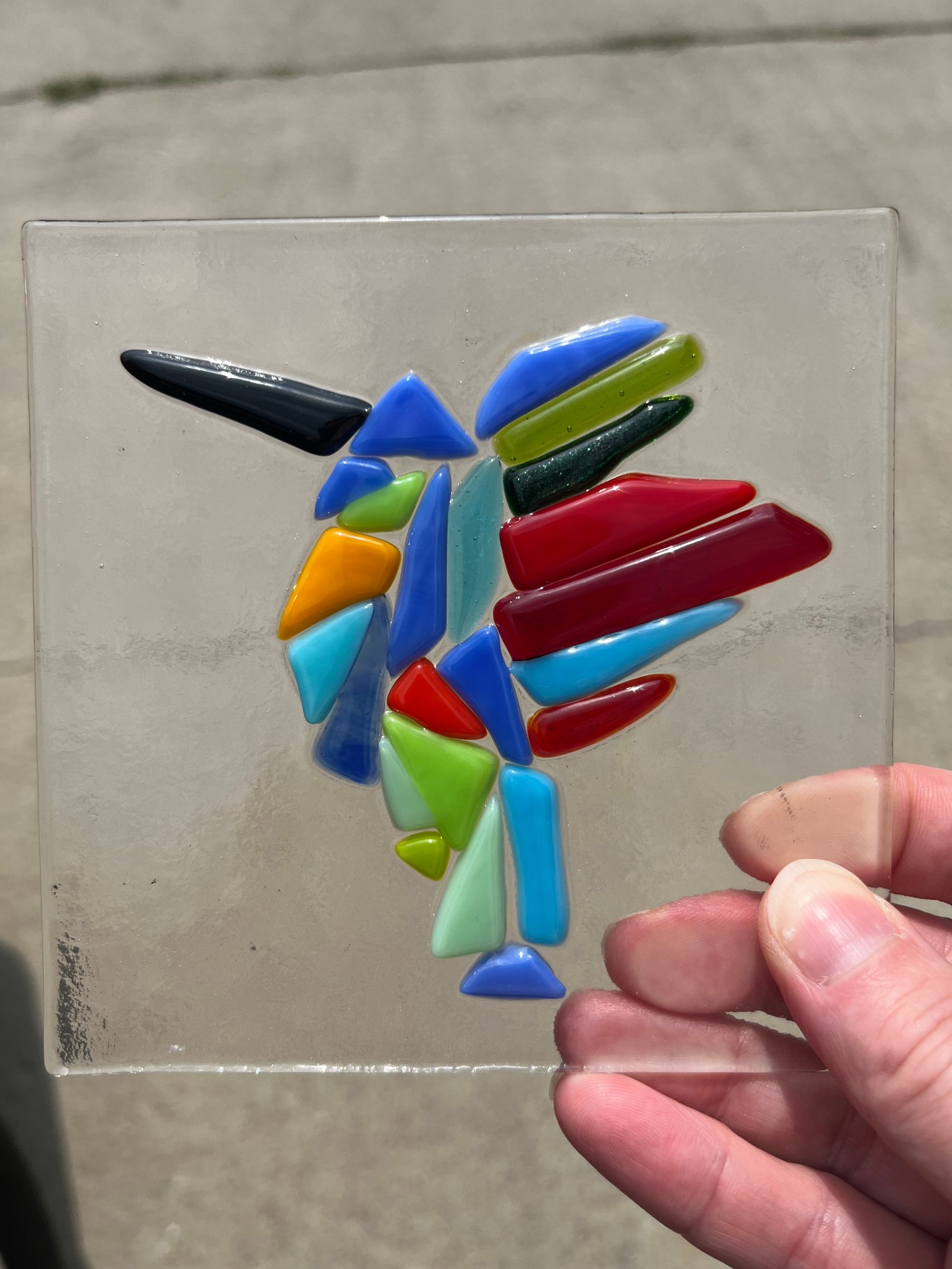 Pieces of different colored glass are fused on a clear glass plate. A right hand is holding the plate.