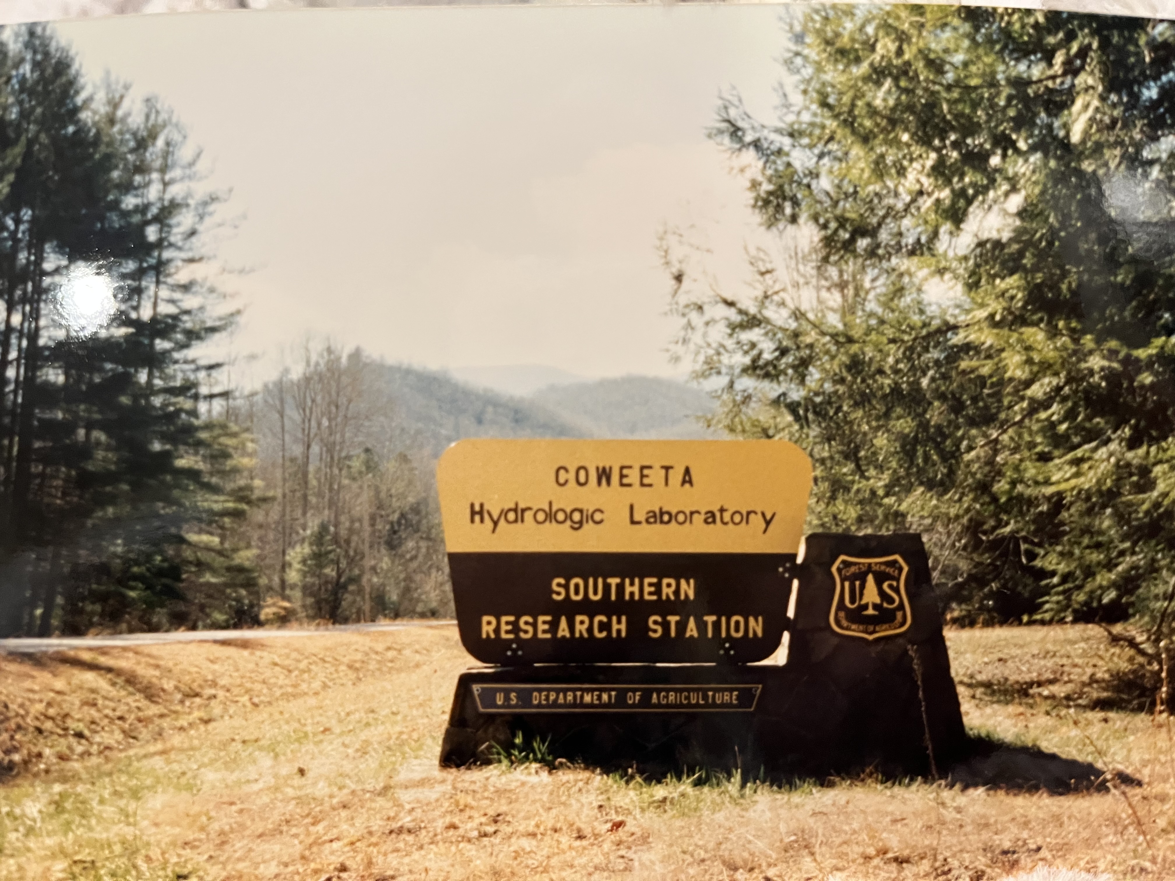 A picture of the entrance sign to Coweeta Hydrologic Laboratory. The picture is somewhat yellowed. There are trees surrounding the sign and rolling mountains in the background.