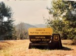 A picture of a sign that says "Coweeta Hydrologic Laboratory Southern Research Station, US Department of Agriculture". There are pine trees around the sign and rolling mountains in the distance. It's a sunny day. The picture is yellowed.