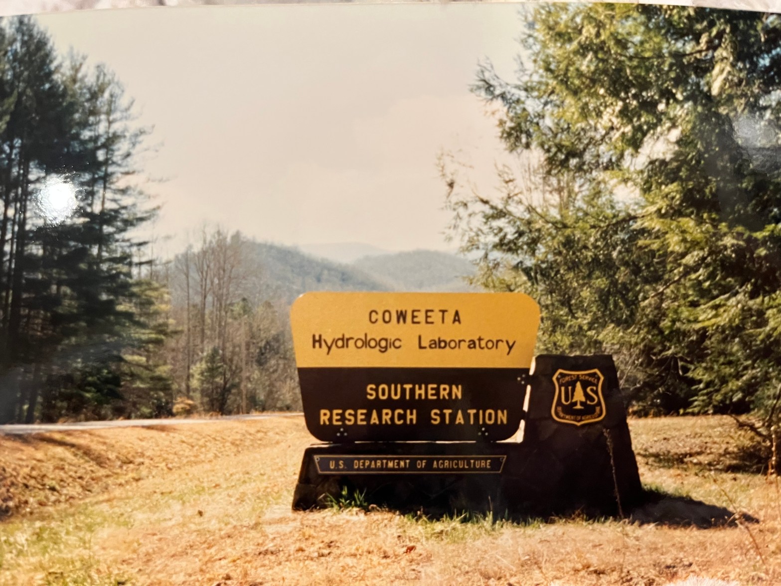 A picture of a sign that says "Coweeta Hydrologic Laboratory Southern Research Station, US Department of Agriculture". There are pine trees around the sign and rolling mountains in the distance. It's a sunny day. The picture is yellowed.