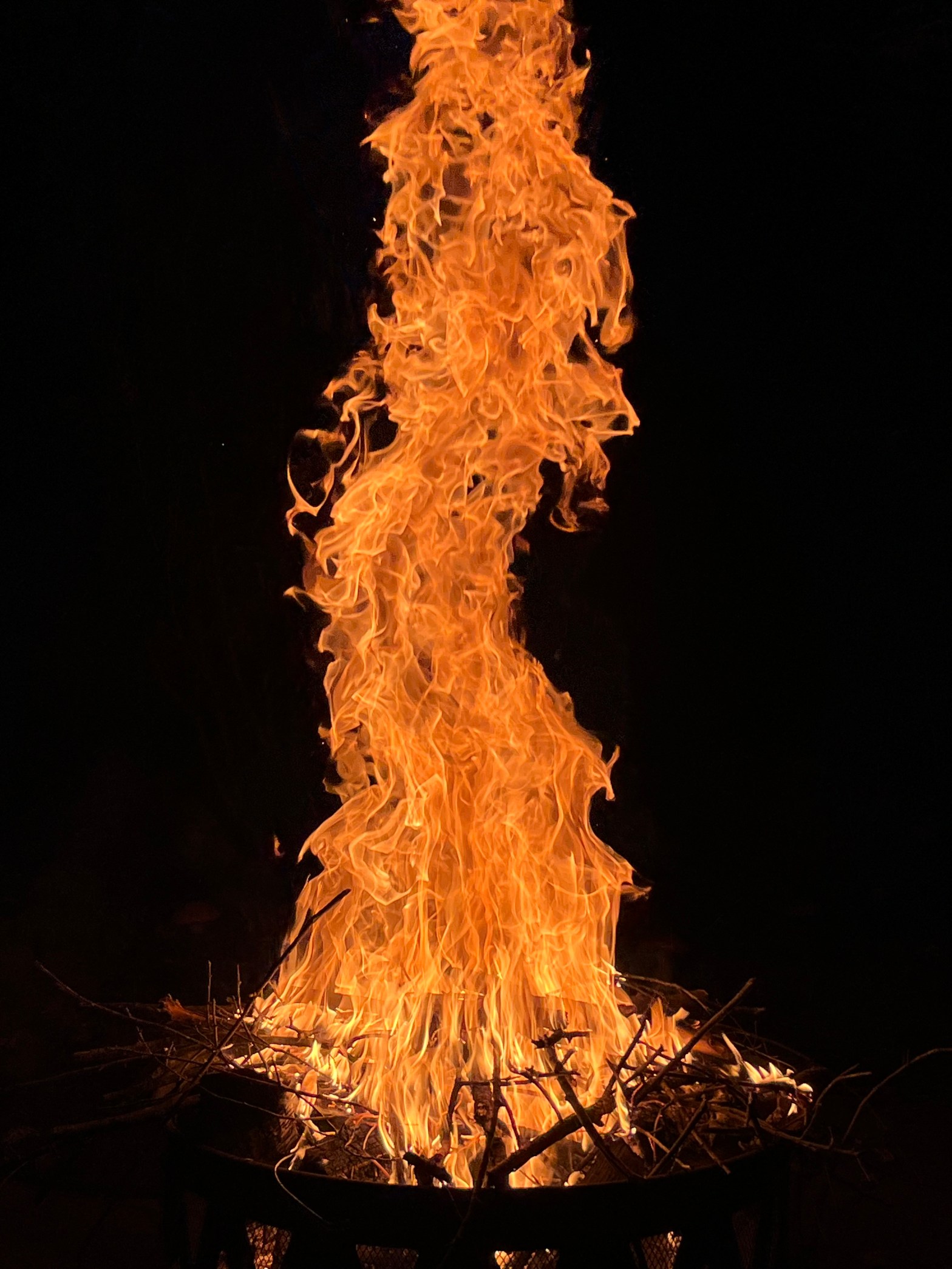 A large fire in a firepit. The fire is about person-sized, with sticks at the bottom. It is bright orange.